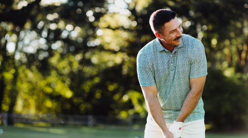 A man wearing a green and white patterned short-sleeve polo shirt and white pants, smiling on a golf course with a blurred green background.
